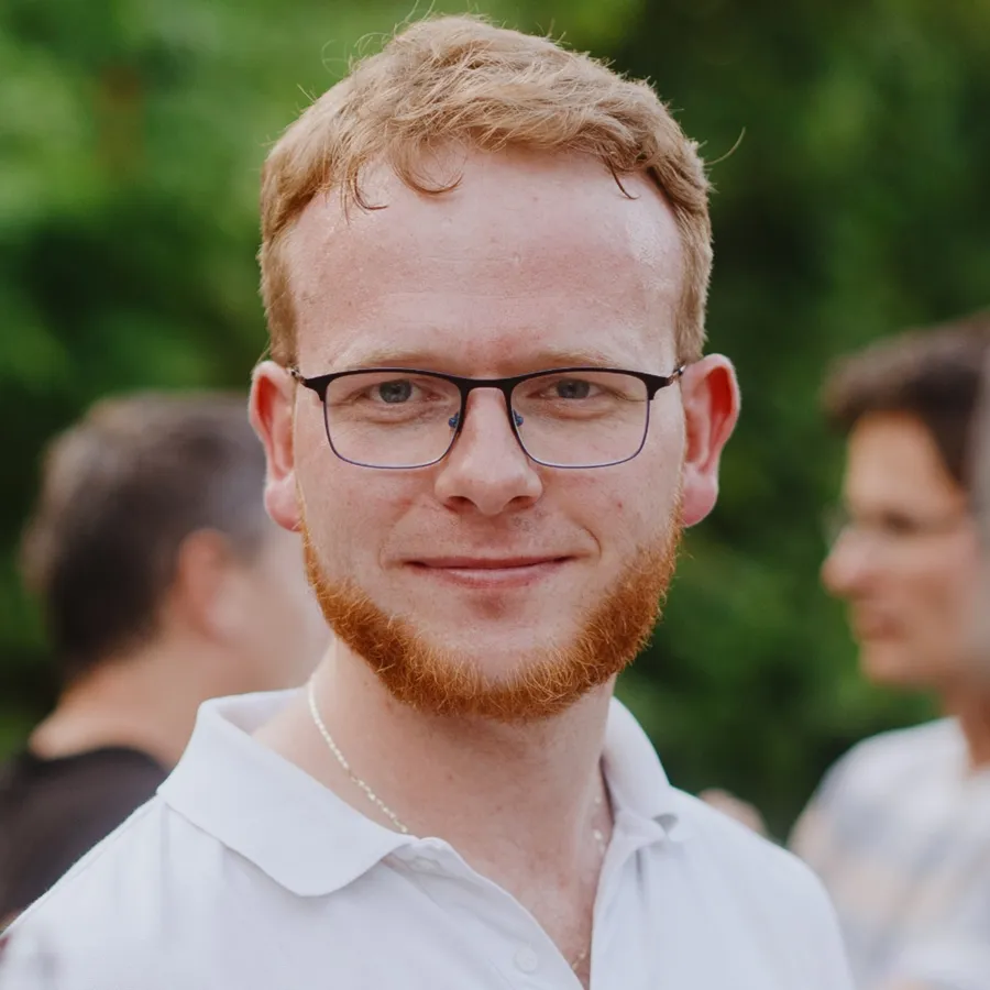 Daniel outdoors in summer, smiling, in a white polo shirt, with friends talking behind him.
